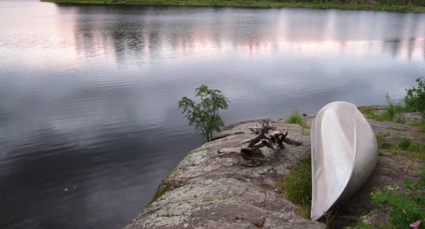 A canoe rests upside down on a rocky shore beside a very calm body of water. 